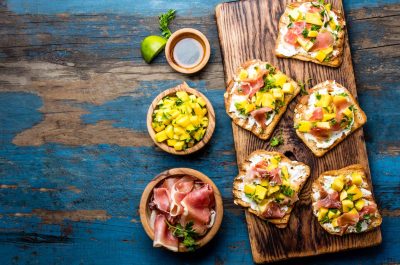 Toasts with cream cheese, ham jamon serrano and mango served on wooden board with red wine, blue wooden rustic background, top view, copy space.